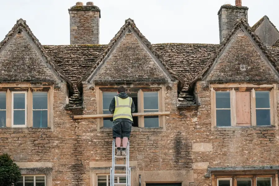 Worker on a ladder repairing a brick house roof under three dormer windows