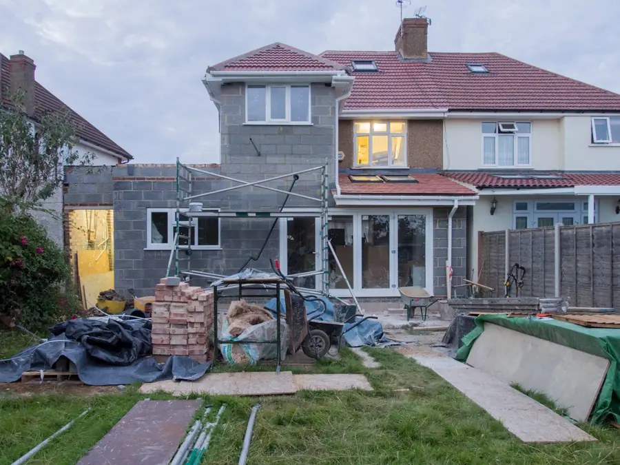 Backyard of a house with scattered construction materials, a wheelbarrow, and a scaffold under a gray sky