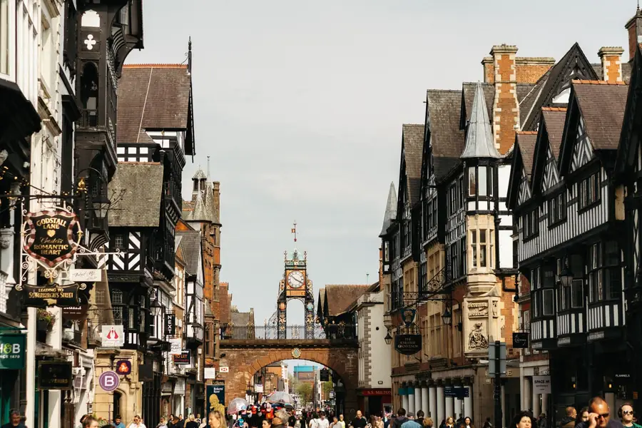 Busy historic street with black-and-white Tudor buildings and a distant clock tower, crowded with pedestrians