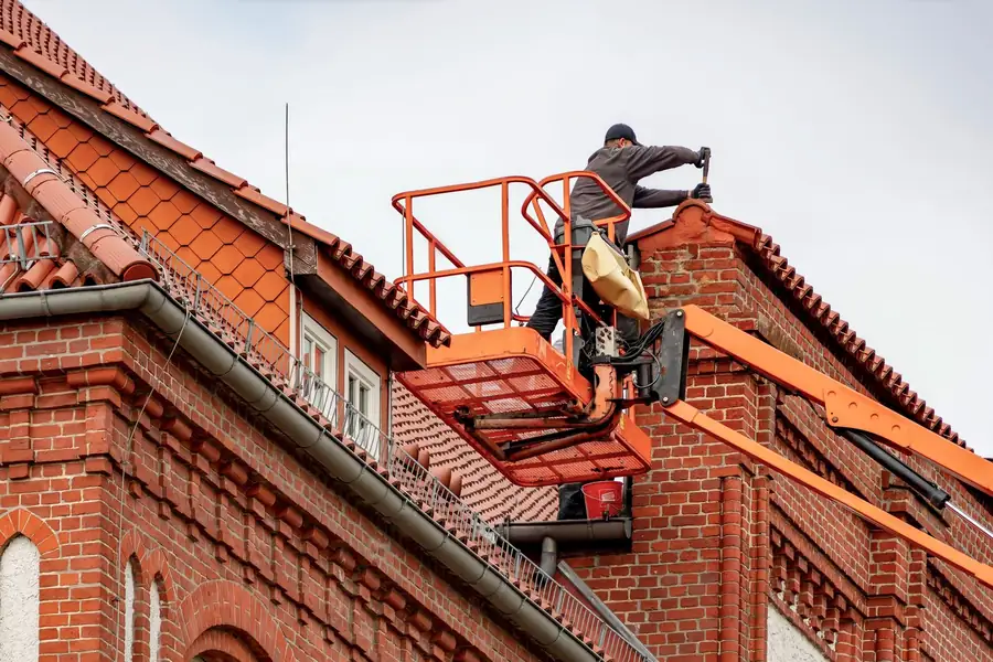 Worker on orange lift repairing a red-tiled roof on a brick building