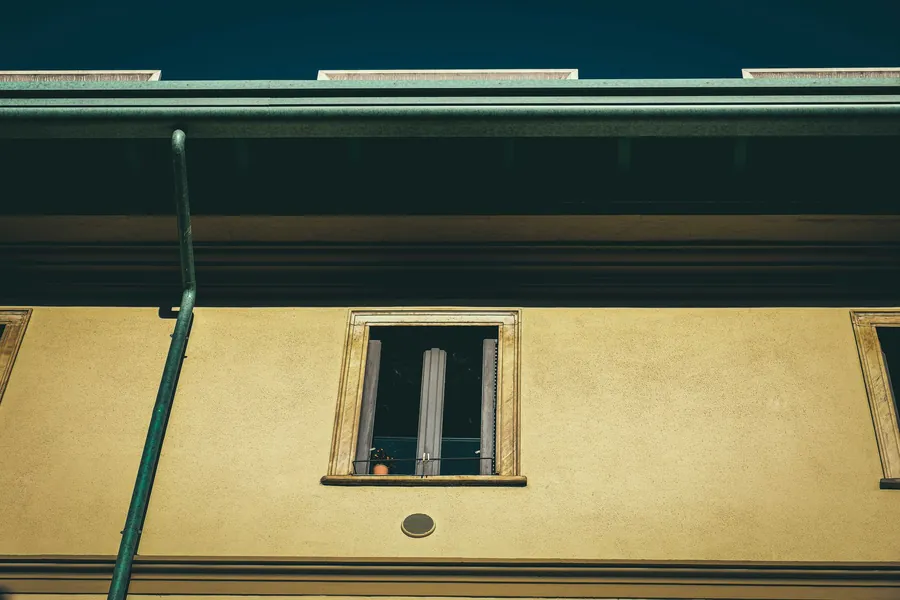 Yellow building facade with a narrow open window and a green downspout below the roofline