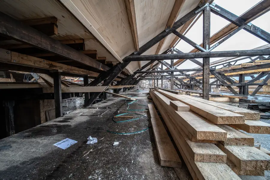 Abandoned industrial warehouse with wooden beams, broken roof trusses, and stacked lumber on a concrete floor