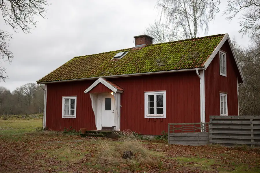 Red wooden cottage with white trim and mossy roof in a rural field