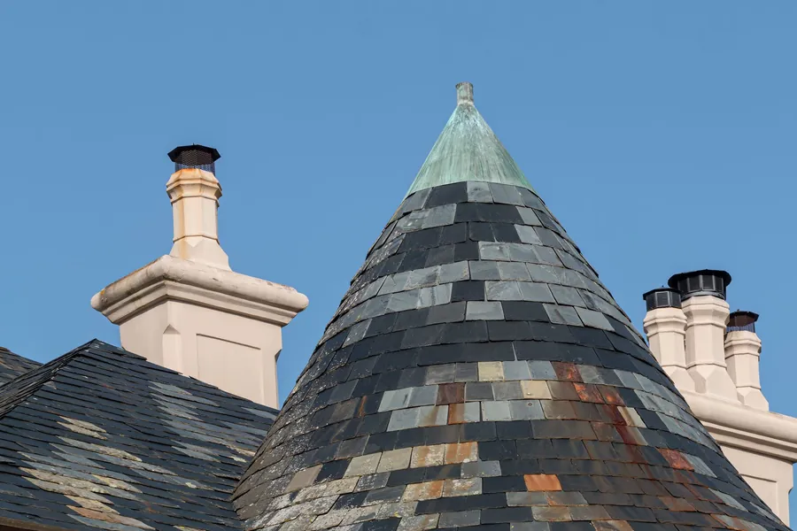 Slate roofs and chimneys against a blue sky, with a conical turret topped in green patina.