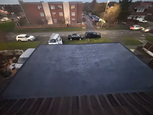 An elevated view of a flat, dark grey roof surface, with residential buildings and parked cars visible in the background.