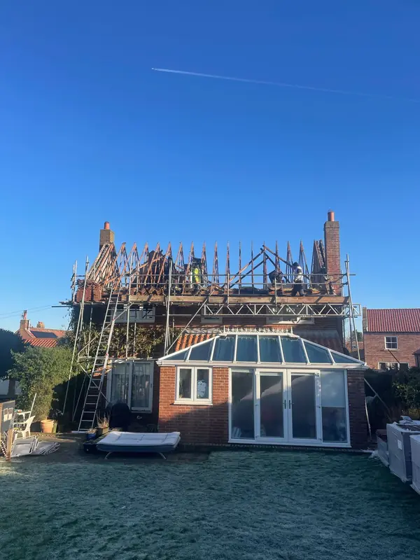 A residential house under construction, with scaffolding around the roof level, seen from a backyard on a sunny day.