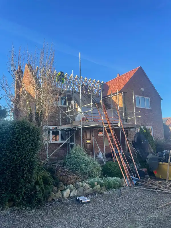 A brick house under construction, featuring scaffolding around the exterior and a bright red tiled roof under a blue sky.
