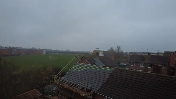 An aerial view of a building with a partially tiled roof, neighboring houses, and a green field under a cloudy sky.