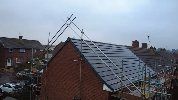 Roofers installing shingles on a house roof. One hammers, the other works in the background. Blue sky overhead.