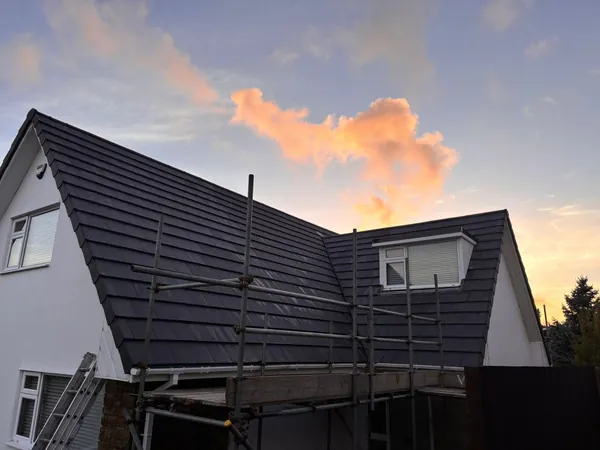 Scaffolding surrounds the roof and white exterior walls of a house at sunset, with dark grey roof tiles and two windows.
