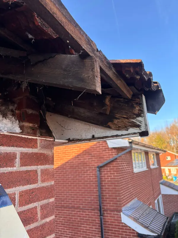 Damaged wooden roof eaves and exposed rafters on a brick house against a blue sky.