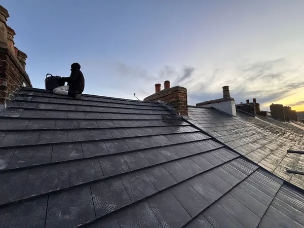 A person sits on a dark, slate-tiled pitched roof against a twilight sky with multiple brick chimneys visible.