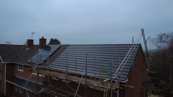 A house roof under construction, featuring new dark tiles, partially installed solar panels, and surrounding scaffolding.