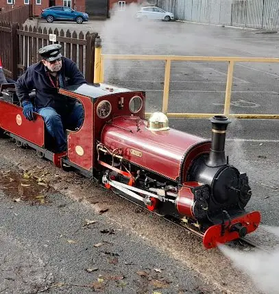 Man driving a small red steam locomotive on a street, with white steam billowing from the front.