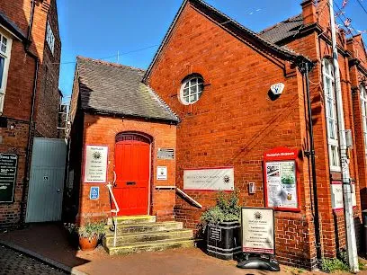 Brick church entrance with red door, steps, and informational signs on a sunny street