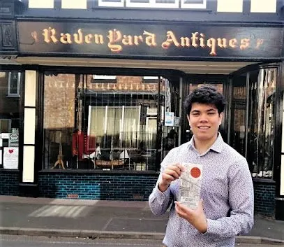 Person holding a book outside a storefront labeled “Raven Gard Antiques.”
