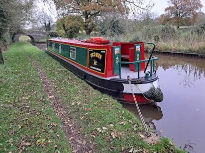 A green and red narrowboat moored beside a canal towpath with autumn trees and a bridge in the distance